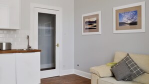 A kitchen with white cabinets, a wooden countertop, and a stainless steel toaster. There is a door with a frosted glass panel, a beige sofa with a plaid pillow, and two framed pictures on the wall. - 1 bedroom accommodation in Millport, Isle of Cumbrae (Millport, Isle of Cumbrae)