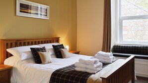 A neatly made bed with white linens and a plaid blanket, a bedside table with a lamp, and a window with curtains. - 1 bedroom accommodation in Millport, Isle of Cumbrae (Millport, Isle of Cumbrae)