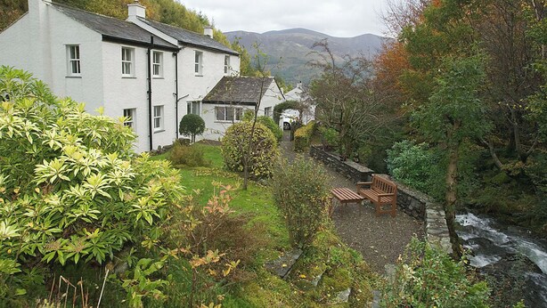 A white building with multiple windows, a stone pathway, and a bench by a stream.