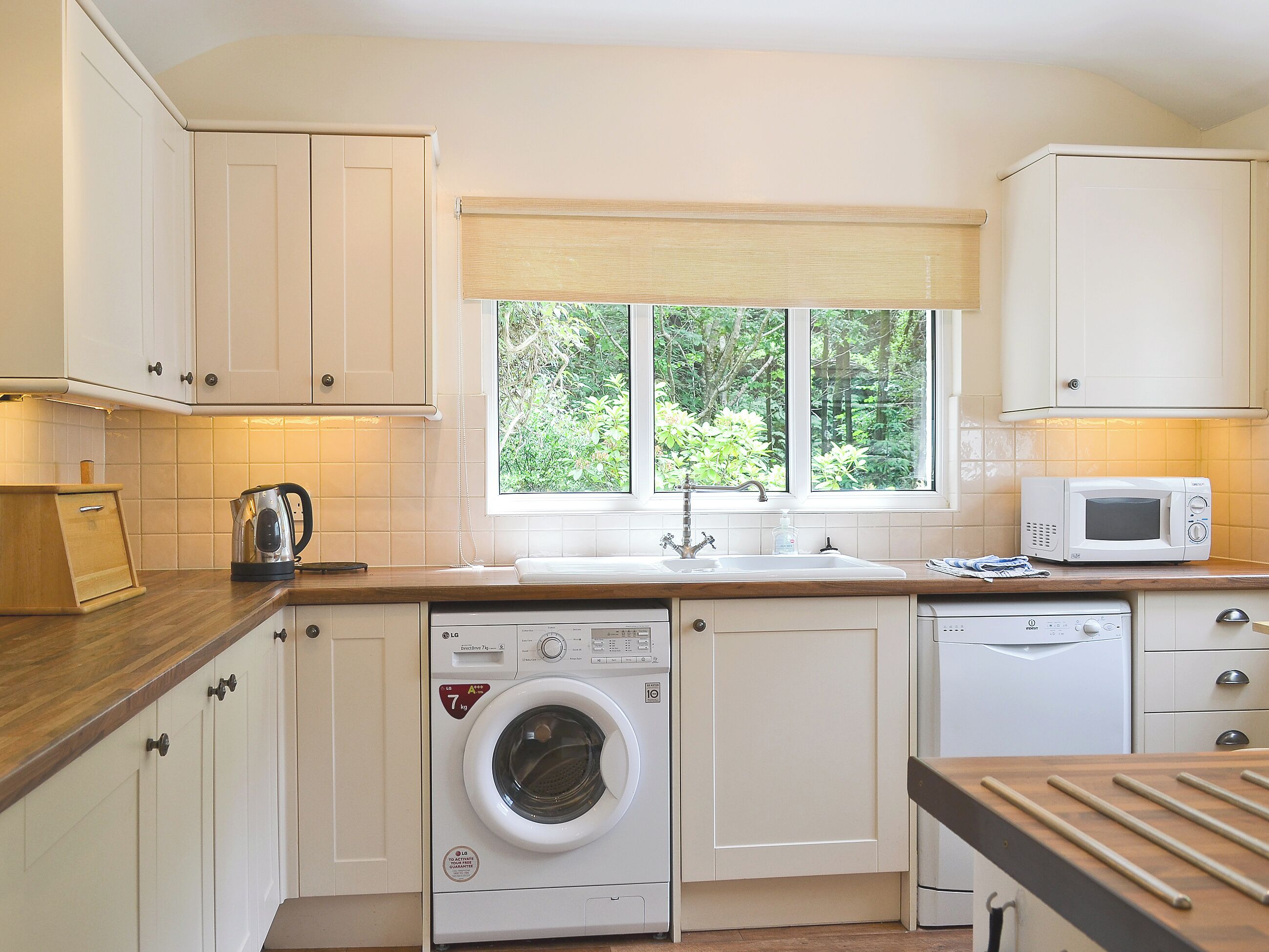 A compact kitchen with white cabinets, a washing machine, a microwave, and a window with a view of greenery.
