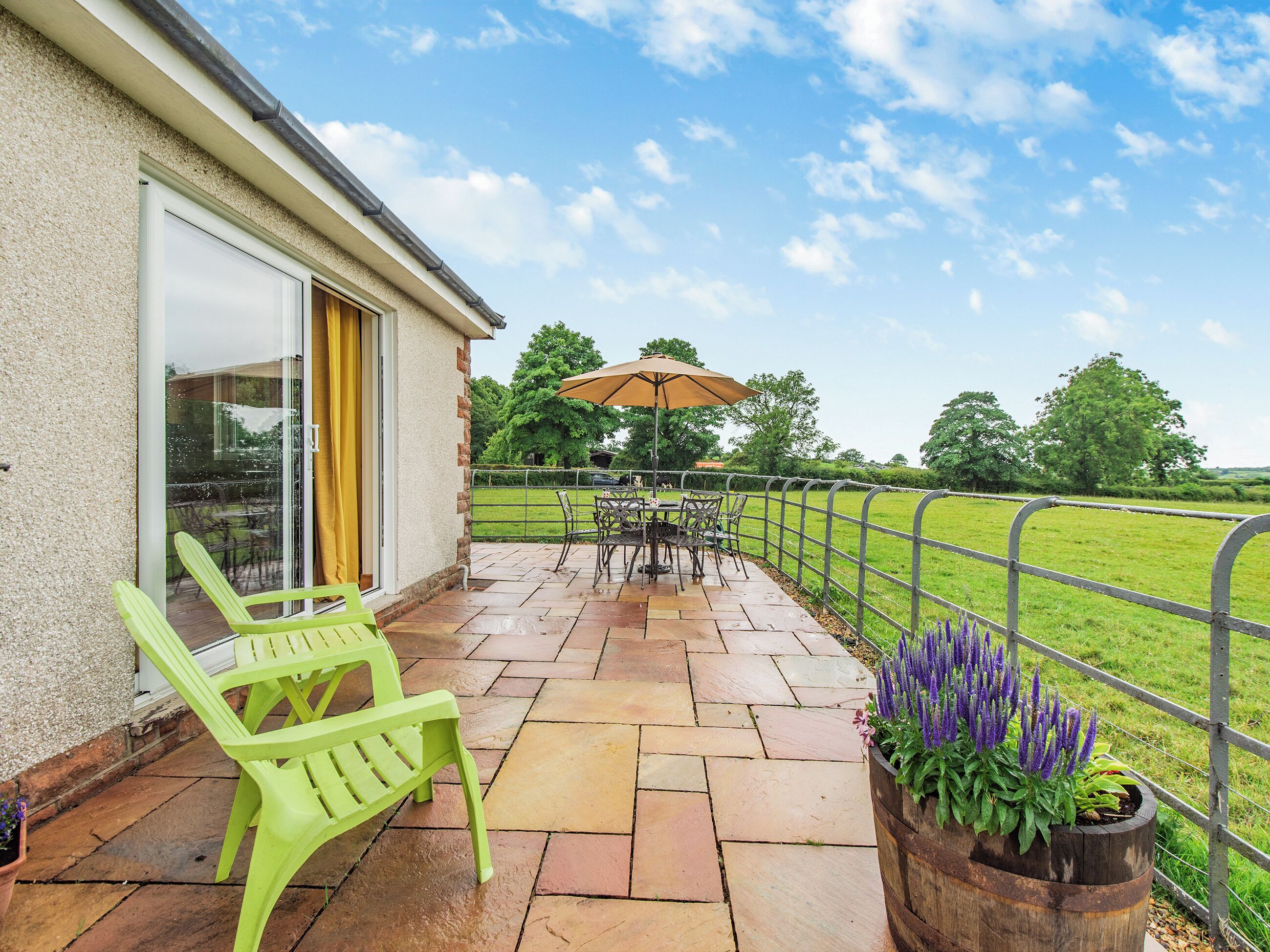 A covered patio with a table and chairs, a green chair, a wooden planter with purple flowers, and a view of a green field.