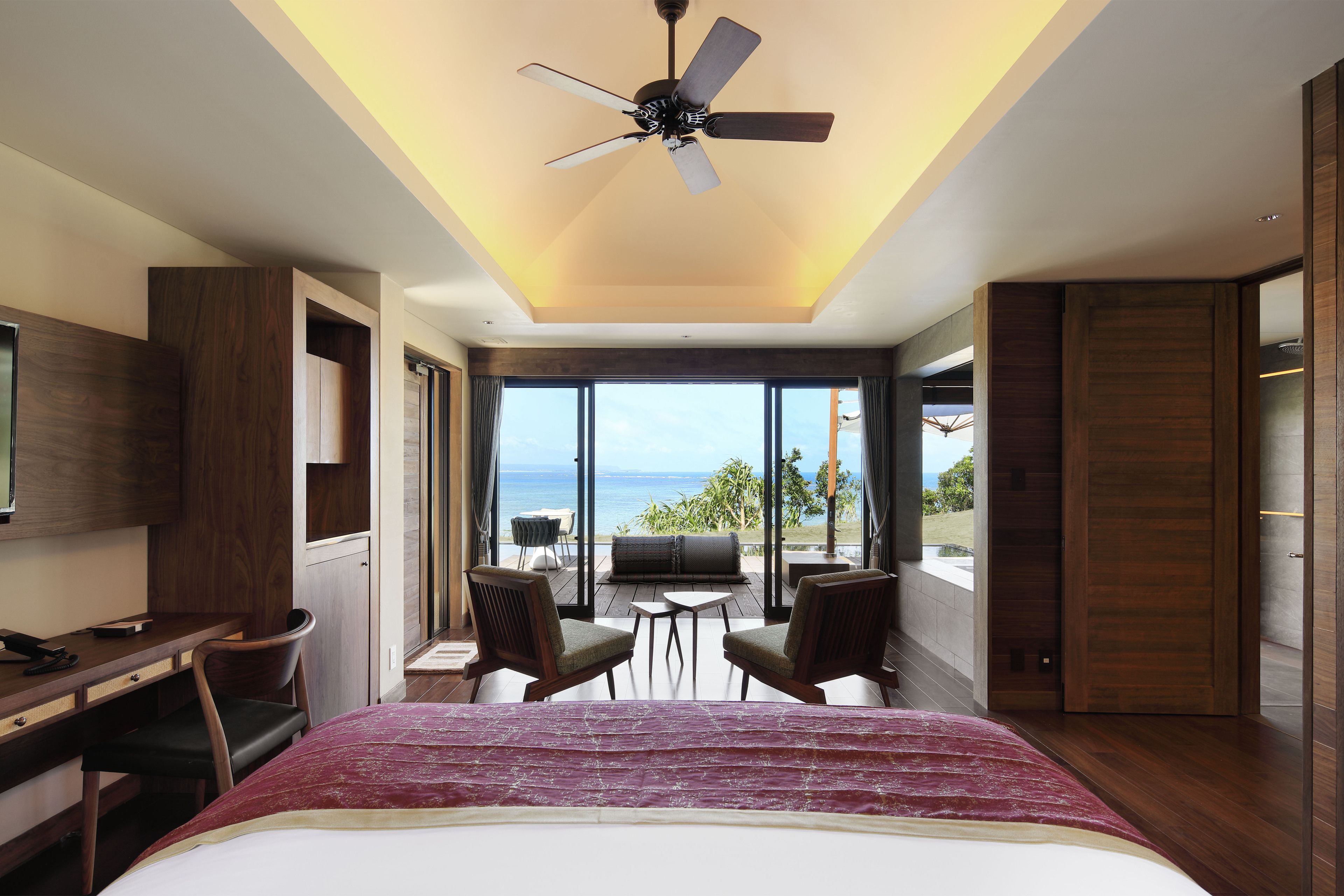 Una habitación de hotel moderna con cama, escritorio, sillas y un ventilador de techo. Cuenta con un amplio ventanal con vista al océano.