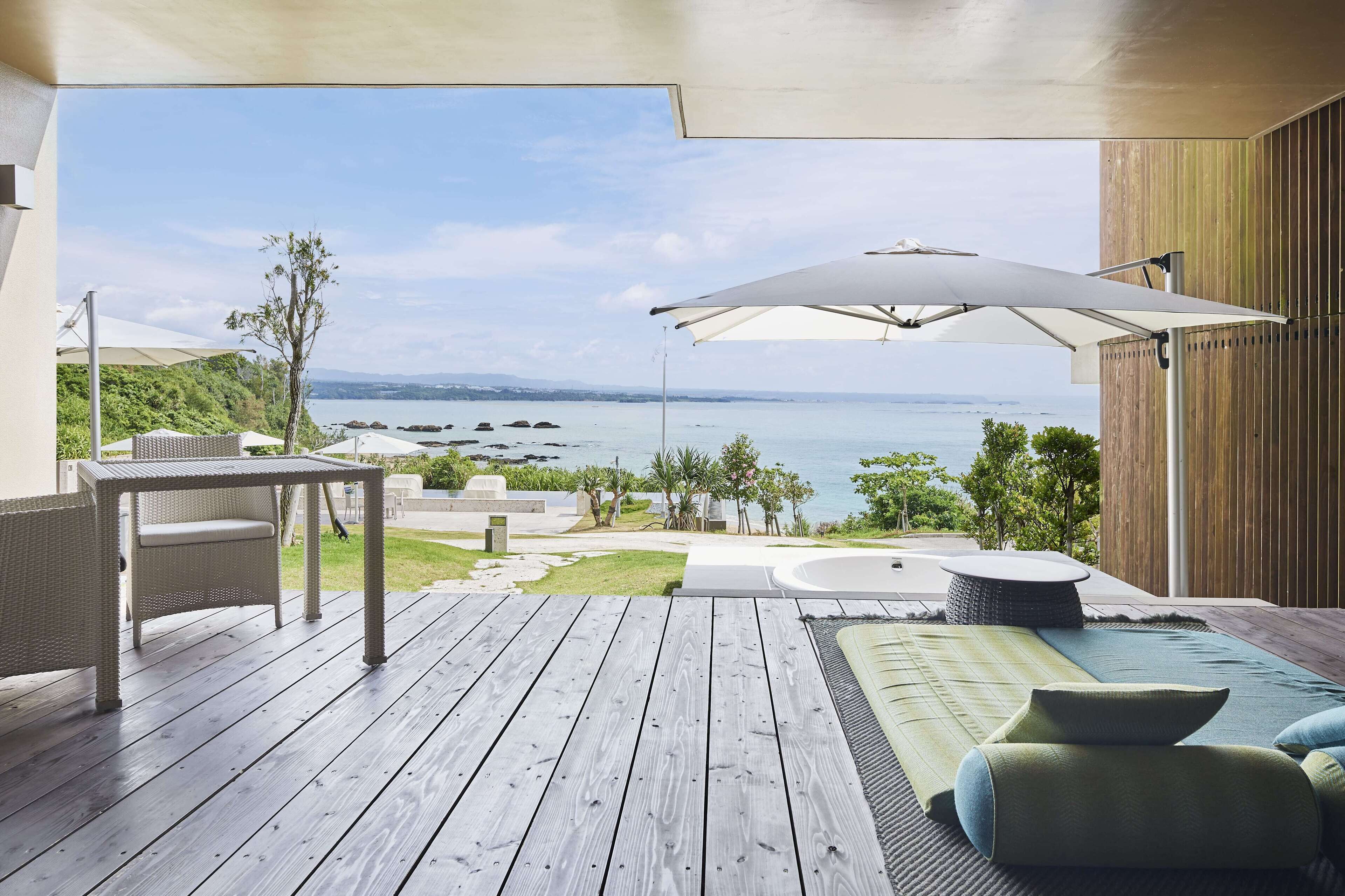 Eine Holzterrasse mit Tisch und Stühlen, einem großen Sonnenschirm und Blick aufs Meer.