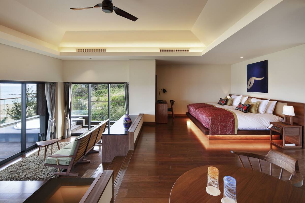 Een moderne hotelkamer met een groot bed, een bureau, een stoel en een bank. Er is een groot raam met gordijnen, een plafondventilator en een schilderij aan de muur.