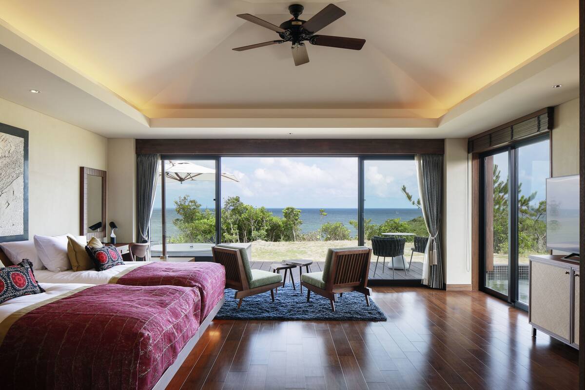 Een ruime slaapkamer met een groot bed, twee stoelen, een klein tafeltje en een televisie. Er is een plafondventilator, een groot raam met uitzicht op de oceaan en een balkon met buitenmeubilair.