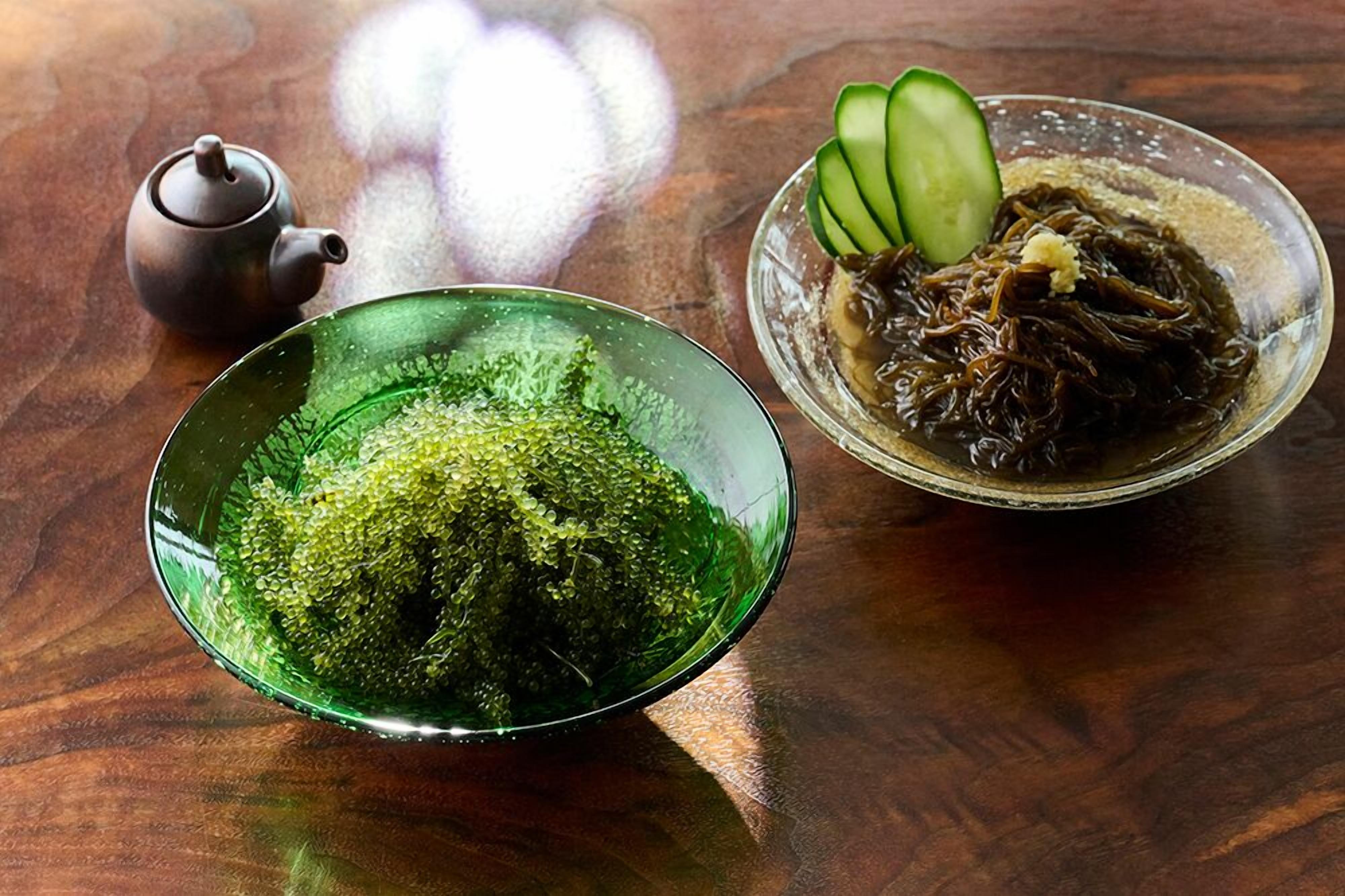 A green bowl of green tea, a teapot, and a bowl of dark soup with cucumber slices on a wooden table.