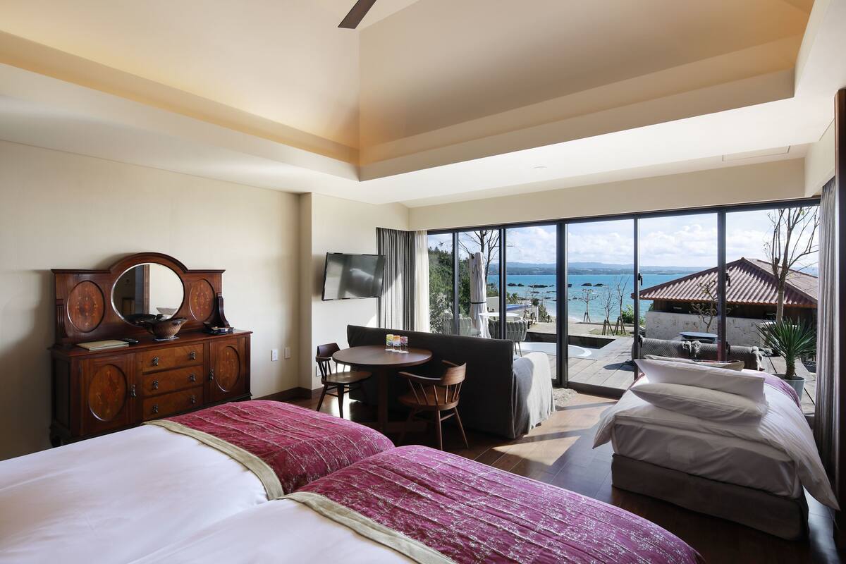 Een slaapkamer met een groot bed, een houten dressoir, een ronde tafel met stoelen en uitzicht op zee door grote ramen.
