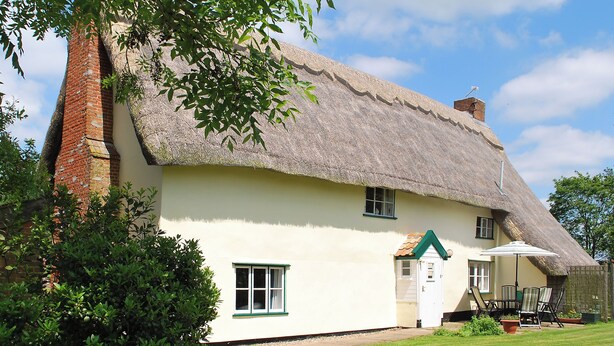 Ein traditionelles Haus mit Strohdach und Backsteinschornstein, ein kleiner Anbau und eine Rasenfläche mit Gartenmöbeln.