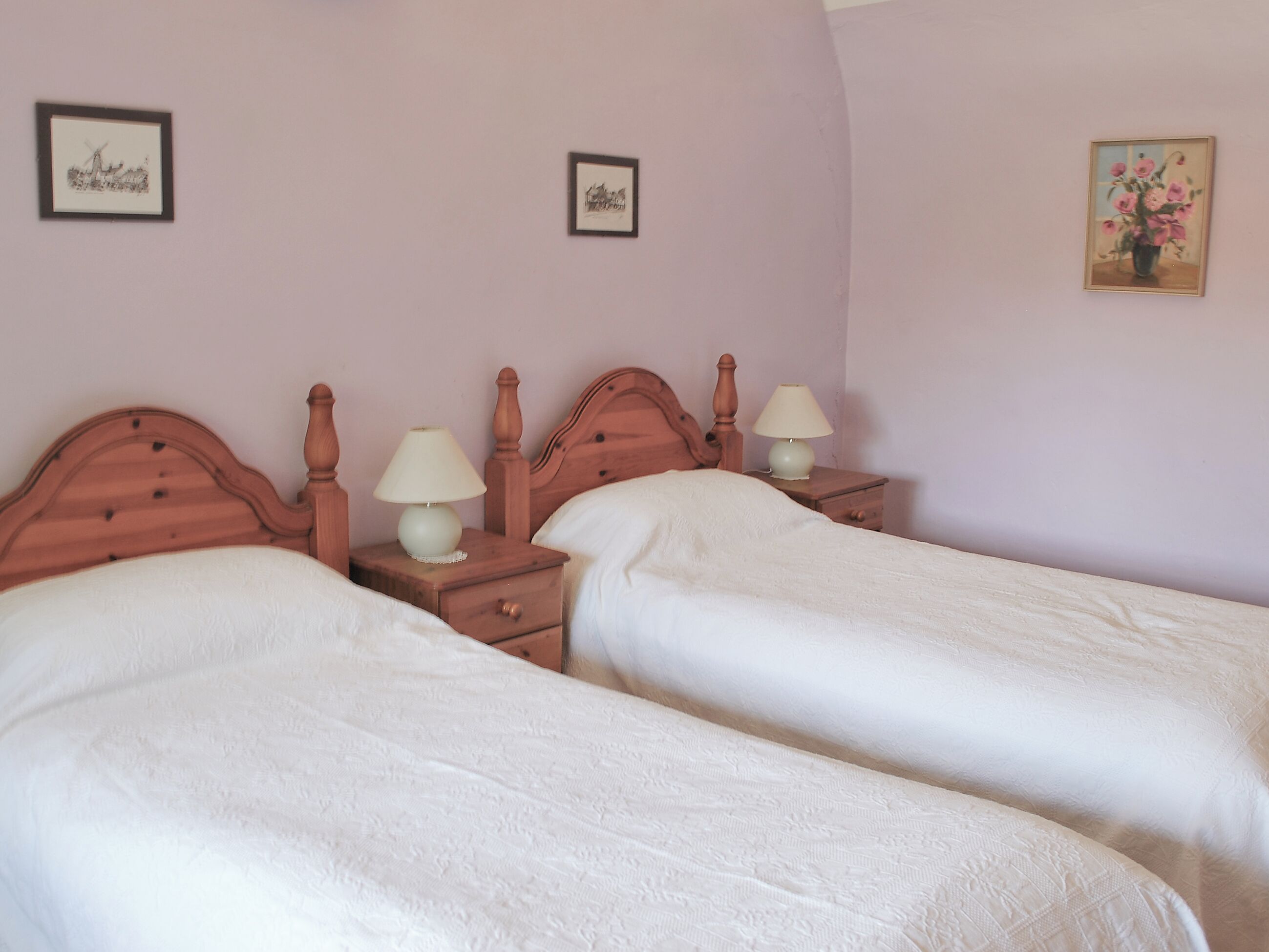 A bedroom with two single beds, wooden headboards, bedside tables with lamps, and framed pictures on the walls.
