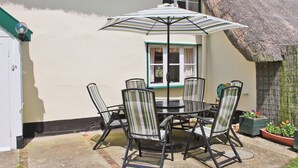 Ein runder Tisch mit sechs Stühlen und einem gestreiften Sonnenschirm auf einer Terrasse mit Strohdach und weißen Wänden.