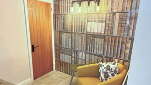 A room with a wooden door, a bookshelf filled with books, a yellow armchair with a floral pillow, and a small table. - A traditional Lakeland cottage situated in the heart of Portinscale village. (Portinscale, near Keswick)