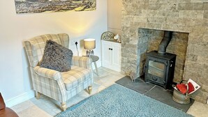 A cozy living room with a plaid armchair, a lamp, a fireplace, and a scenic painting on the wall. - A traditional Lakeland cottage situated in the heart of Portinscale village. (Portinscale, near Keswick)