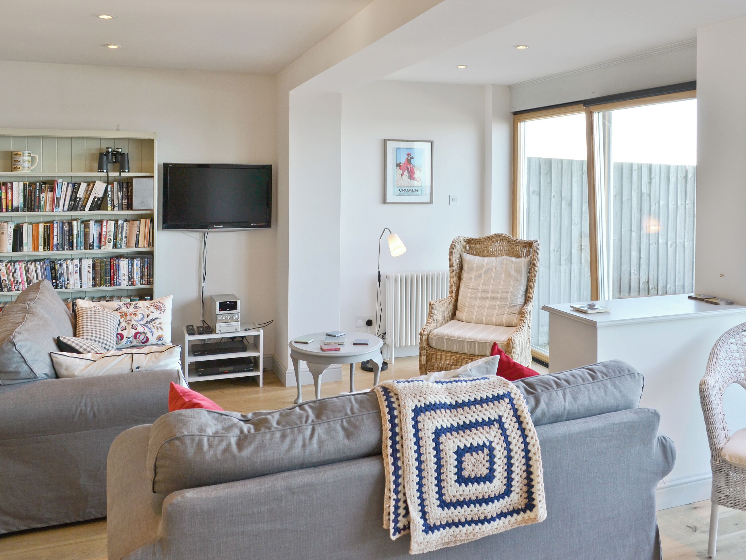 Een woonkamer met een grijze bank, een boekenkast, een televisie, een rieten fauteuil en een raam met jaloezieën.