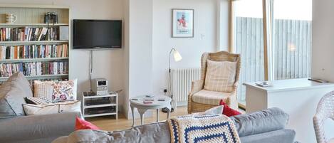 Een woonkamer met een grijze bank, een boekenkast, een televisie, een rieten fauteuil en een raam met jaloezieën.