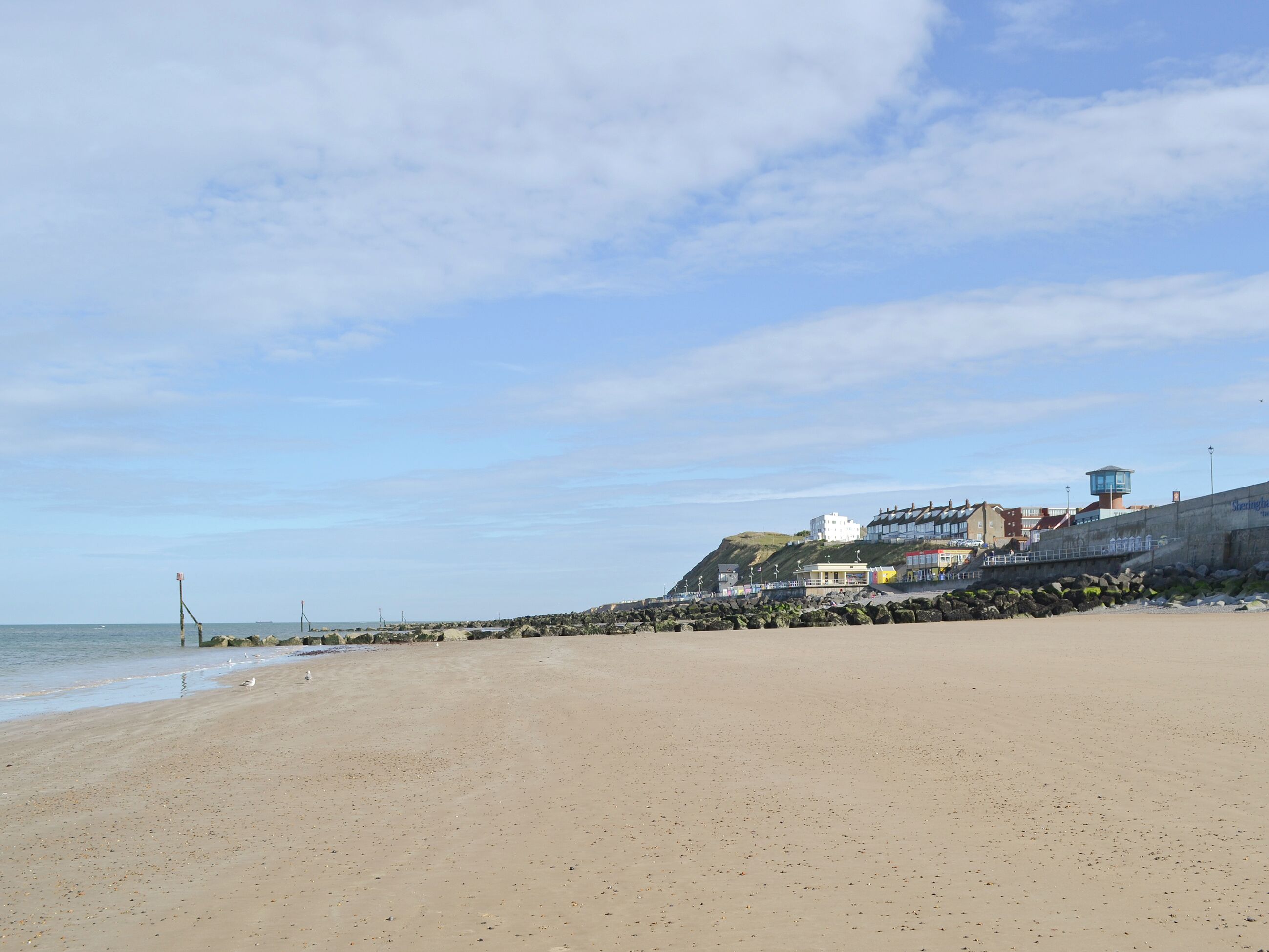 Ein weiter Sandstrand mit einem steinernen Wellenbrecher und Gebäuden im Hintergrund.