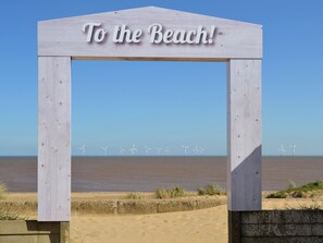 Une arche en bois portant l’inscription « À la plage ! » se dresse devant une plage, avec des éoliennes en arrière-plan.