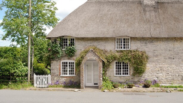 Ein traditionelles Steinhaus mit Strohdach, weißem Zaun und einem kleinen Garten mit violetten Blumen.