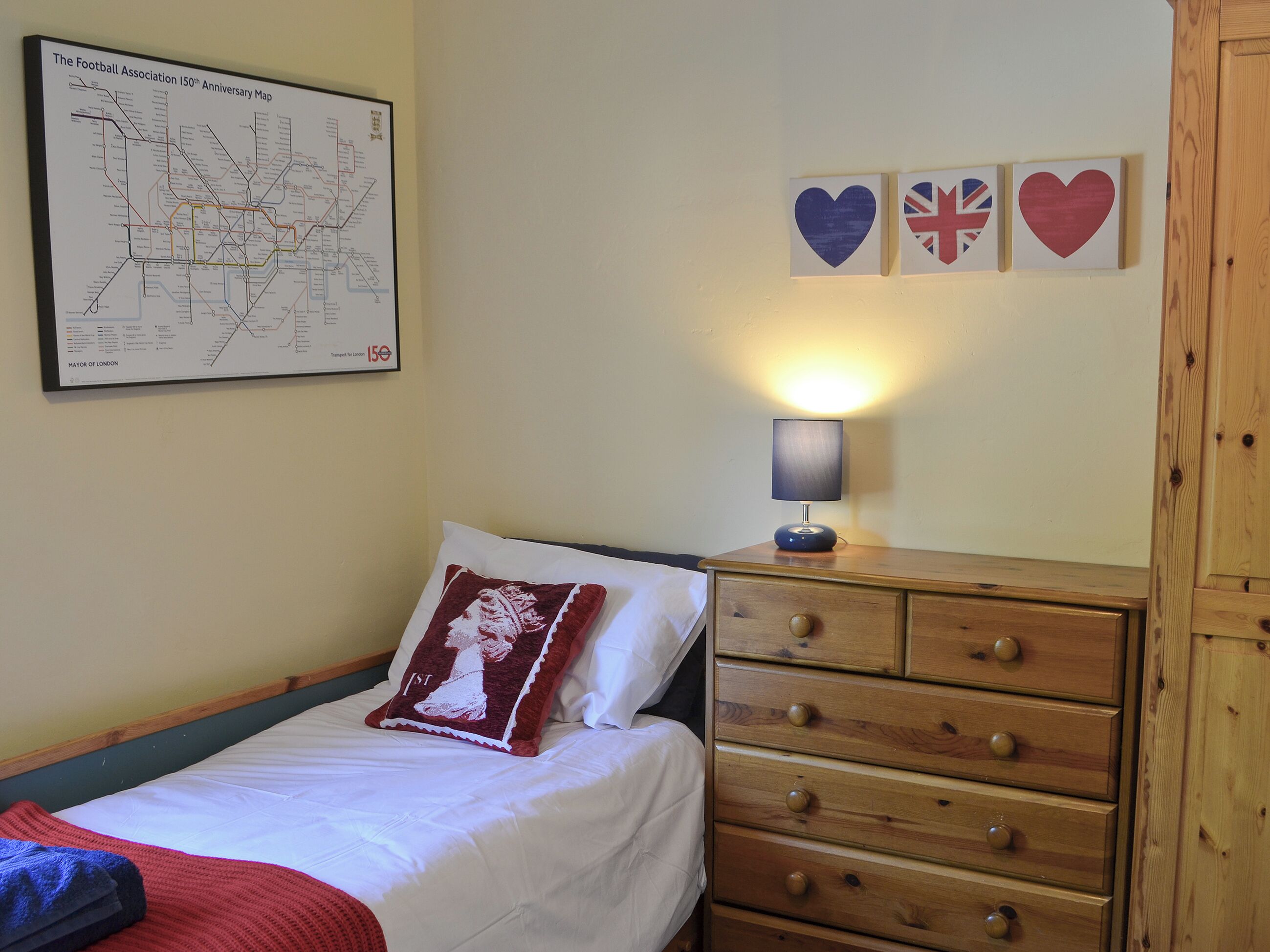 Un lit bien fait, avec un oreiller rouge et blanc, une commode en bois dotée de tiroirs, une carte accrochée au mur et trois décorations en forme de cœur ornées de drapeaux.