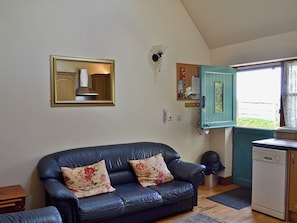 A living room with a dark blue sofa, a wooden cabinet, a wall-mounted light fixture, and a window with a view of the outside. - Jasmine Cottage - 30503 (Beddingham, near Lewes)