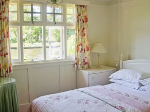 A bedroom with a bed, a nightstand, a lamp, a window with curtains, and a view of greenery outside.