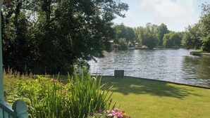 A lakeside view with a wooden fence, a grassy lawn, and a wooden planter with flowers. - 3 bedroom accommodation in Hoveton, near Wroxham, Norwich (Hoveton, near Wroxham, Norwich)