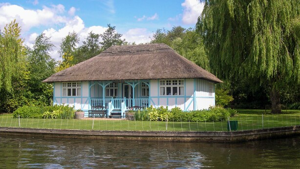 A thatched-roof house with a blue and white exterior, situated by a calm body of water, surrounded by lush greenery. - 3 bedroom accommodation in Hoveton, near Wroxham, Norwich (Hoveton, near Wroxham, Norwich)