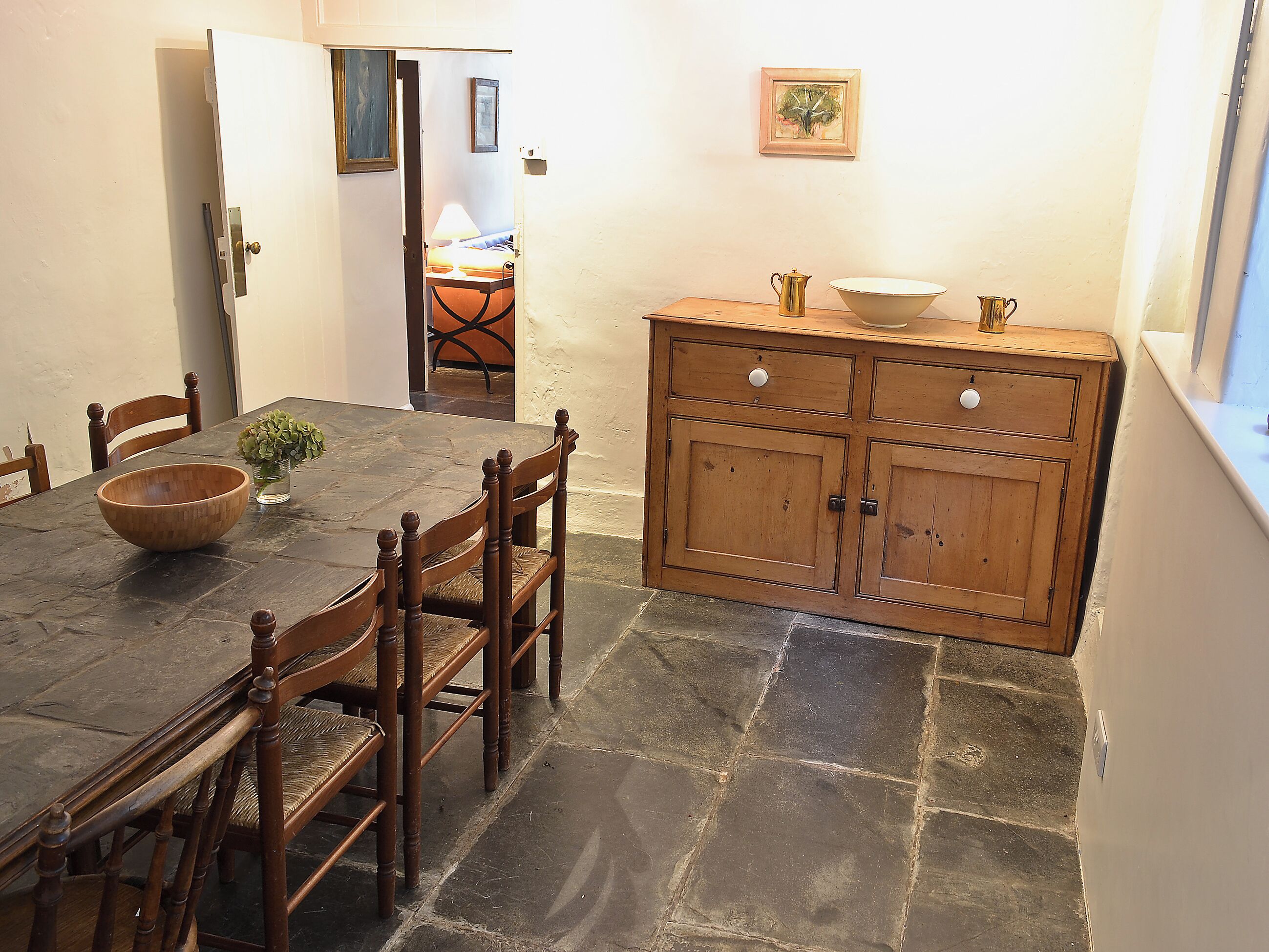 A dining area with a stone floor, wooden table, chairs, and a wooden cabinet with a bowl and jug on top.