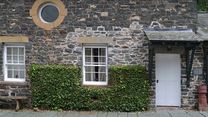 A stone building with two windows and a door, a circular window above the left window, and a hedge in front.