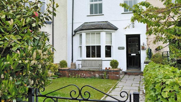Une maison blanche à deux étages, avec une porte d’entrée noire, une baie vitrée et un petit jardin agrémenté d’un portail en métal.