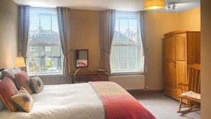 Une chambre à coucher comprenant un lit, une armoire en bois, un fauteuil à bascule, un bureau avec un miroir et de grandes fenêtres avec des rideaux.