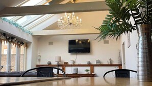Un salon moderne avec une table à manger en bois, deux chaises, un lustre, un téléviseur à écran plat et une grande plante.