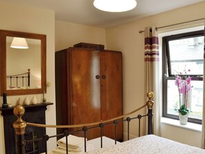 Ein Schlafzimmer mit einem Bett, einem Holzschrank, einem Spiegel, einem Fenster mit Vorhängen und einer Vase mit Blumen.
