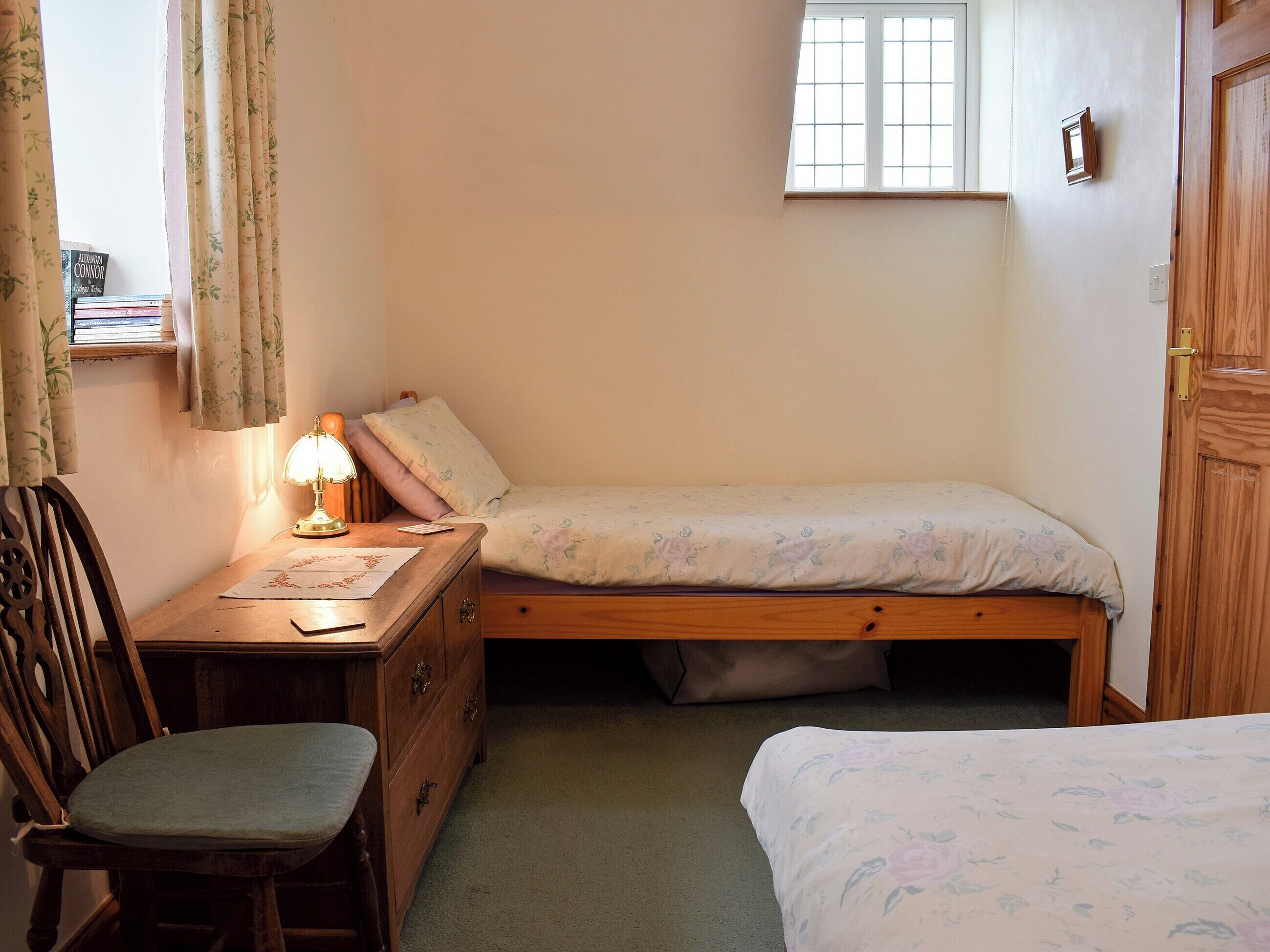 Un dormitorio pequeño y acogedor con una cama individual, una mesita de noche de madera, un escritorio con lámpara, una silla y una ventana con cortinas.