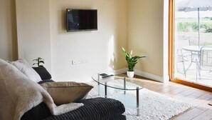 Ein Wohnzimmer mit einem an der Wand montierten Flachbildfernseher, einem Glastisch, einer Couch mit Überwurfdecke, einer Topfpflanze und Blick auf eine Terrasse mit Gartenmöbeln.