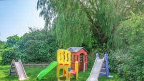 Un terrain de jeux avec des toboggans et un petit bâtiment rouge, entouré d’herbe verte et d’arbres.