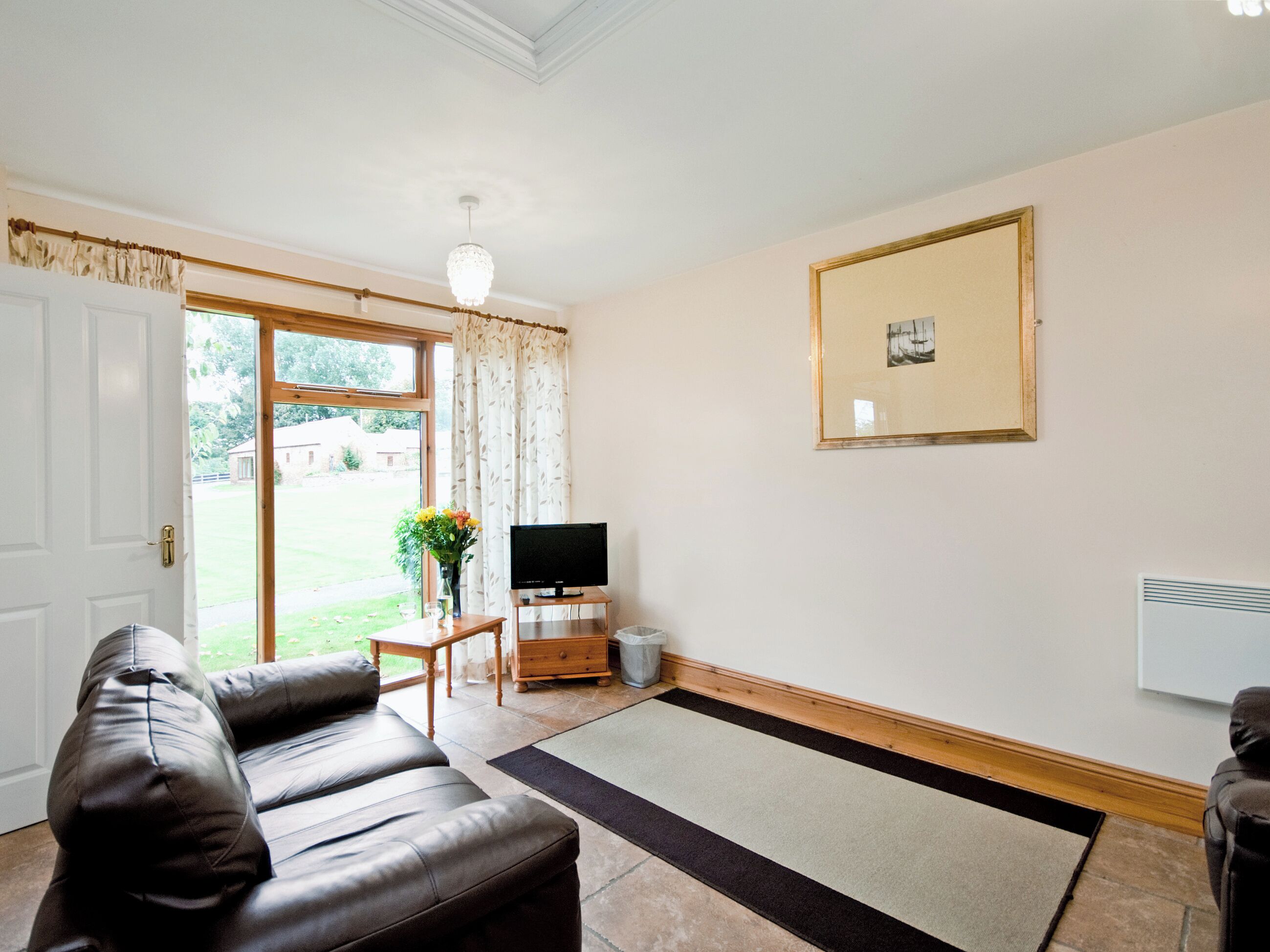 Ein Wohnzimmer mit einer Ledersofa, einem Fernseher auf einem Holzboden, einem gerahmten Bild an der Wand und einer Tür, die in einen Garten führt.