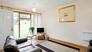 A living room with a leather sofa, a television on a wooden stand, a framed picture on the wall, and a door leading to a garden.