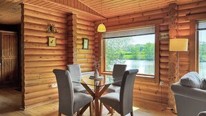 A log cabin interior with a dining table, two chairs, a sofa, and a large window offering a view of a lake. - 2 bedroom accommodation in Hagworthingham (Hagworthingham)