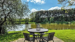 A round metal table with four chairs on a stone patio overlooking a calm lake, surrounded by lush greenery and trees. - 2 bedroom accommodation in Hagworthingham (Hagworthingham)