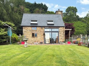Ein Steinhäuschen mit geneigtem Dach, einer großen Glasdoor und einer kleinen Terrasse mit Gartenmöbeln.