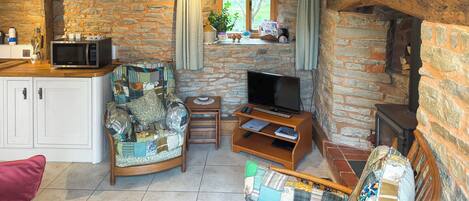 A cozy living room with a stone wall, a wooden ceiling, a television, a small table, and a sofa with a patchwork cover.