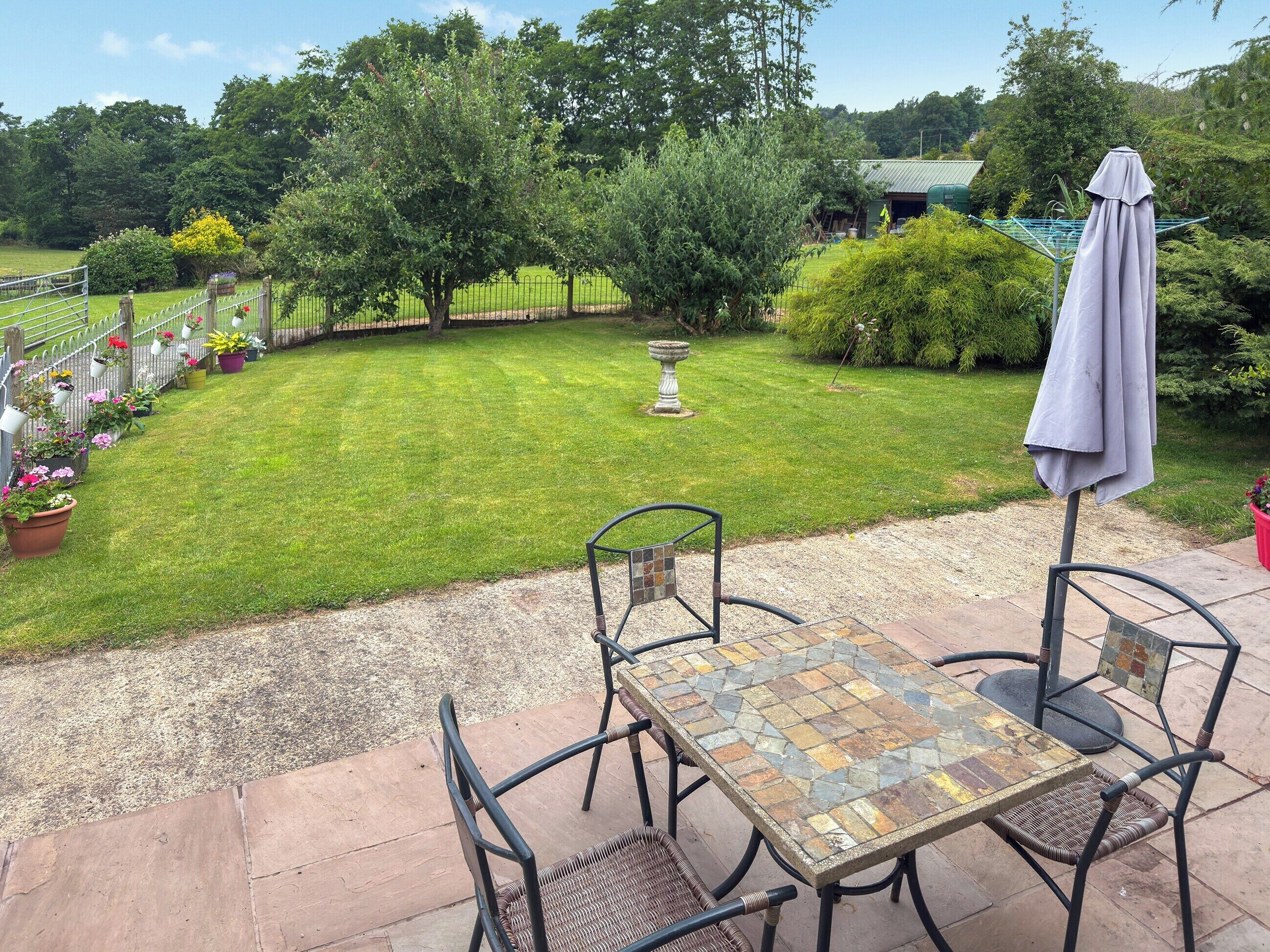 Un espace patio agrémenté d’une table et de chaises décorées de motifs en mosaïque, entouré d’une pelouse et d’un jardin bien entretenus.