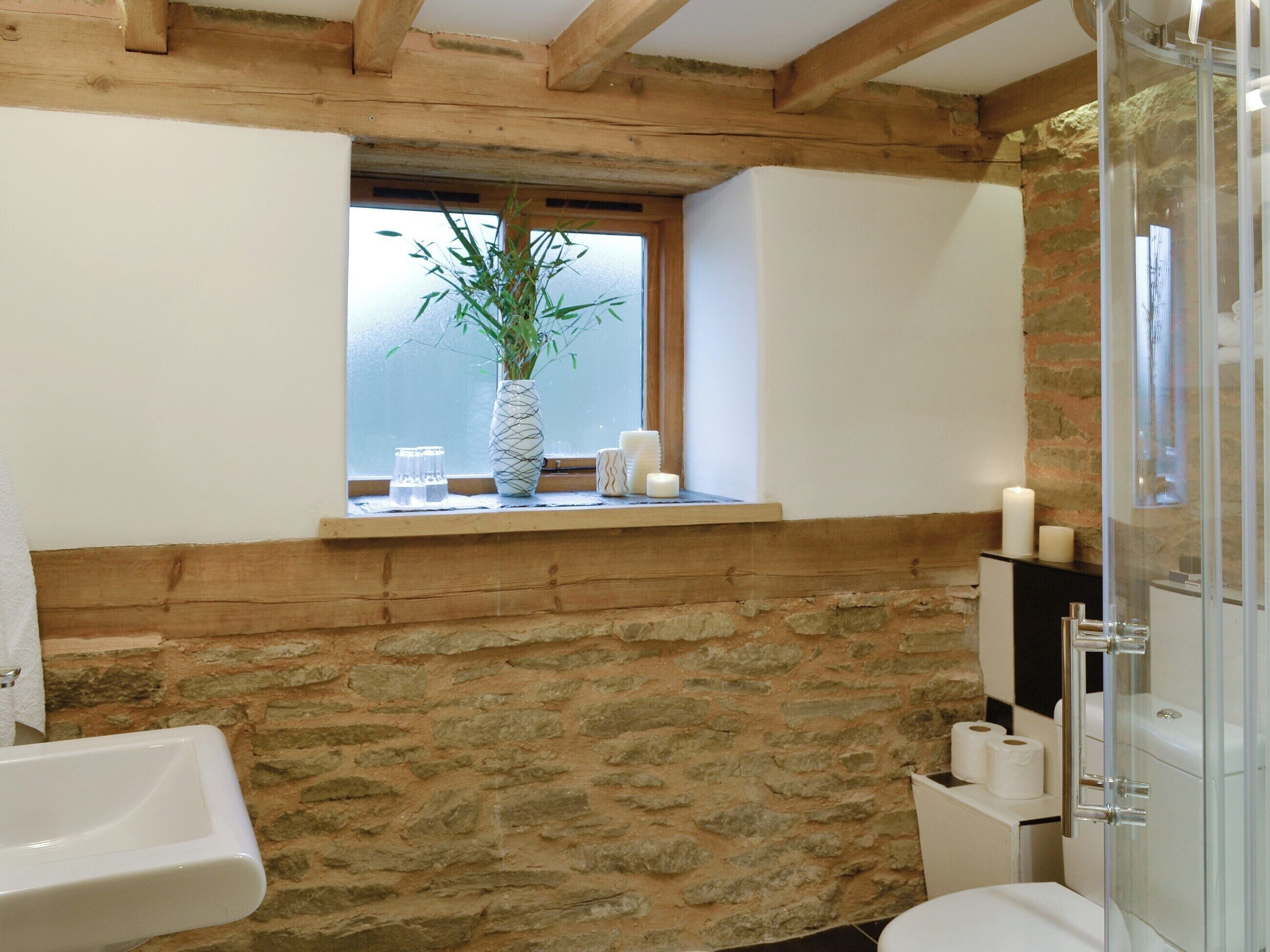 A bathroom with a stone wall, wooden beams, a window with a plant, and a glass shower enclosure.
