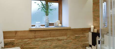 A bathroom with a stone wall, wooden beams, a window with a plant, and a glass shower enclosure.