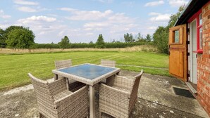 A patio with wicker furniture and a glass table, a brick building with a red window frame, and a grassy field in the background. - Tarrant Cottage -14557 (East Orchard, near Shaftesbury)