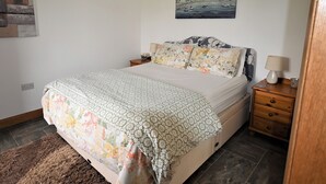 A bedroom with a bed, a nightstand, a lamp, a painting on the wall, and a rug. - Orchard Farm Cottage (Barnby, near Beccles)