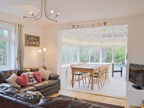A living room with a leather sofa, a dining table with chairs, a television, and a large window with a view of greenery. - 3 bedroom accommodation in Wells-next-the-Sea (Wells-next-the-Sea)