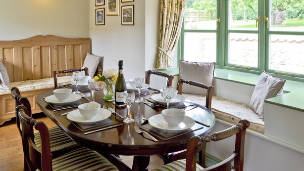 Un comedor con una mesa de madera preparada para cuatro personas, rodeada de sillas, un banco y una ventana con cortinas.