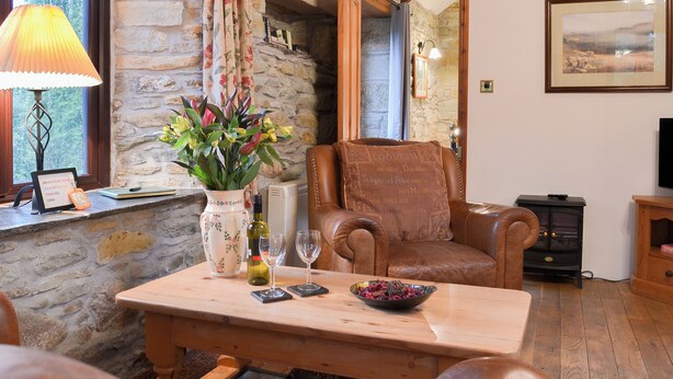 A cozy room with a stone wall, a wooden table with a vase of flowers, a leather armchair, and a television. - 1 bedroom accommodation in Michaelstow, near Camelford (Michaelstow, near Camelford)
