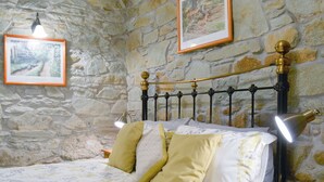 A bed with a stone wall backdrop, a framed picture above it, and a nightstand with a lamp. - 1 bedroom accommodation in Michaelstow, near Camelford (Michaelstow, near Camelford)